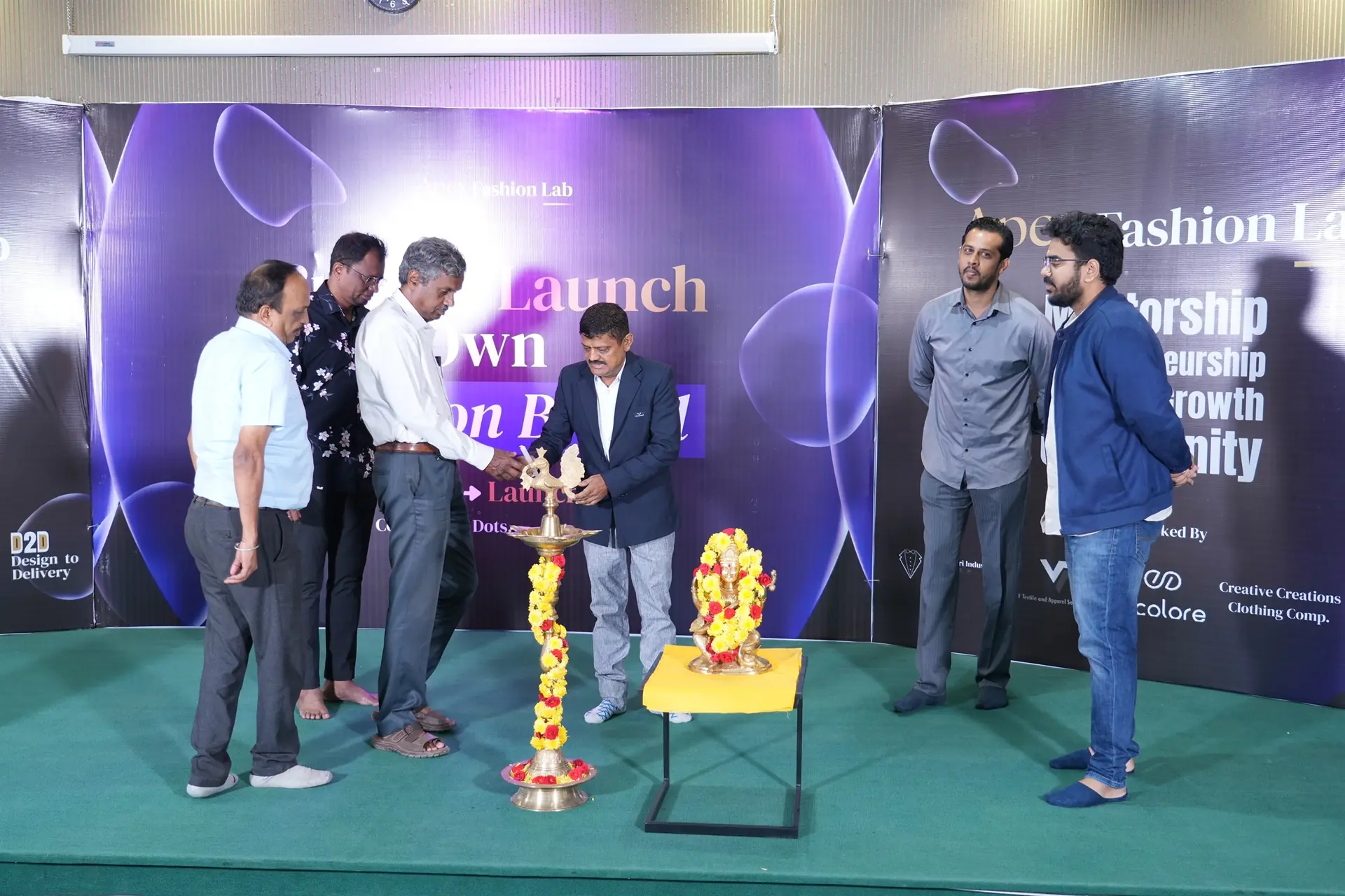 A lamp-lighting ceremony on stage at an Apex Fashion Lab event: one man lights a tall brass lamp hung with marigold garlands while four others stand alongside, in front of a backdrop reading Apex Fashion Lab.
