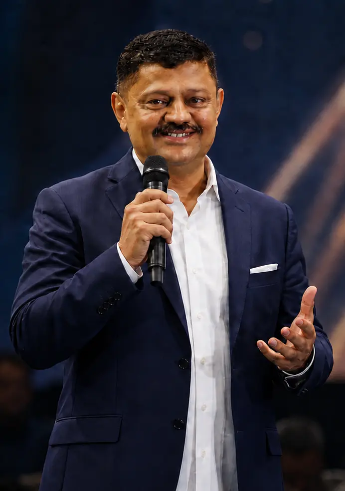 Satyanarayana Sheshagiri speaking on stage with a microphone, addressing an audience